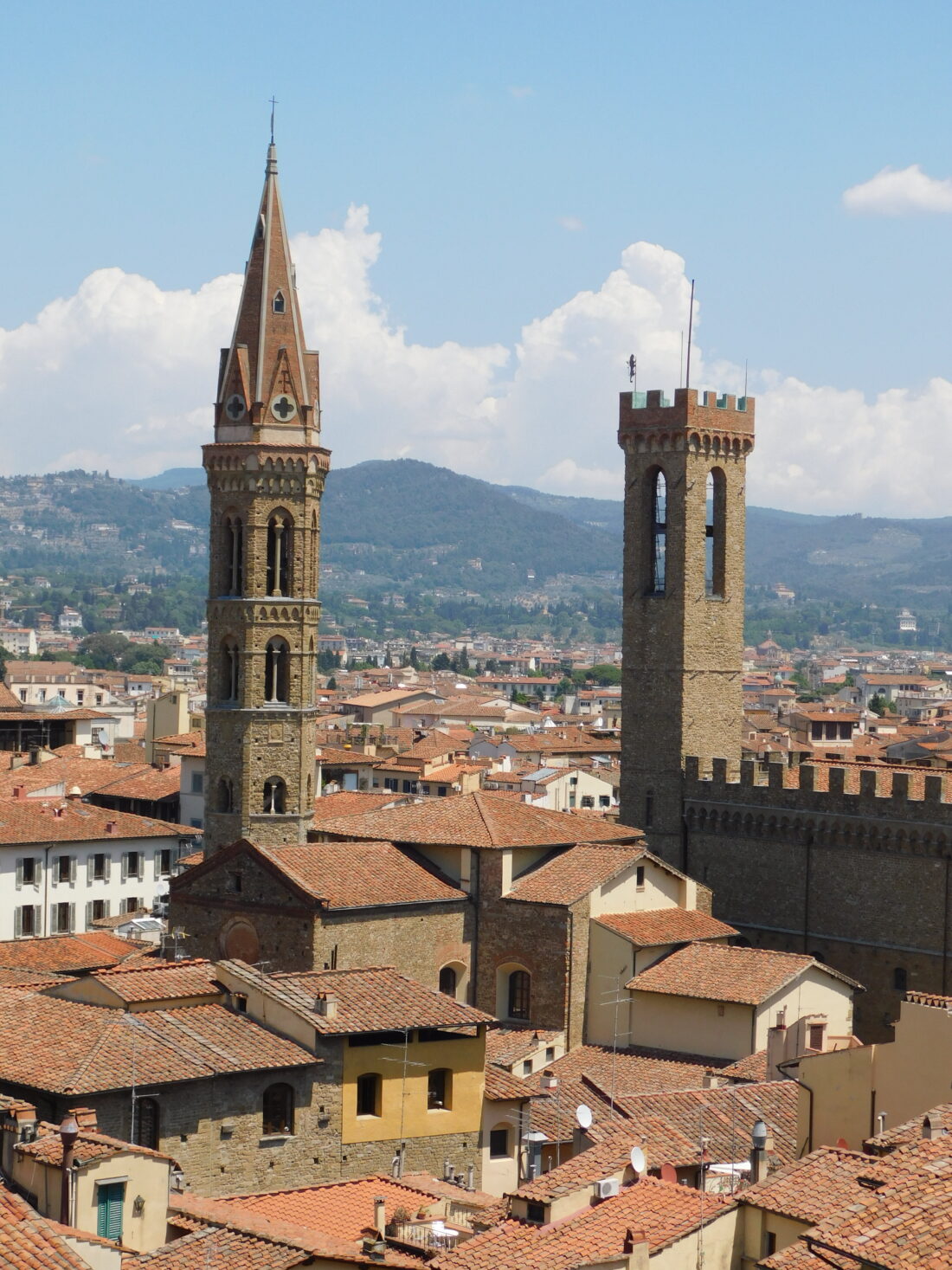 A photograph taken from my tower apartment across the roofs of Florence. Two golden stone towers stick up above all the red tiled roofs. On the left is a pointy one with crosses on it, part of the Badia monastery. On the right is a square one with battlements and big open windows, connected to a fortress with more battlements.