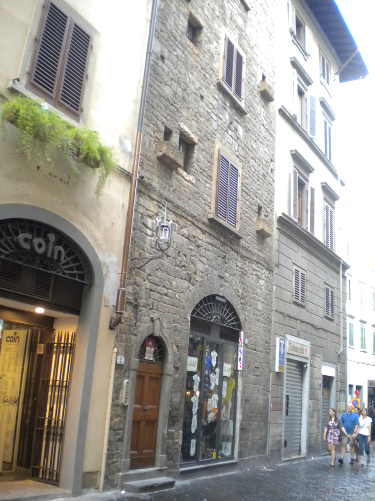 Photo of a street of town buildings, all squeezed together sharing walls with no gaps between. The one on the left is yellow stucco, with an archway and a "Coin" grocery store. The one on the right is yellow stucco above with decorative faux rustic stone facing on the lower floor. In between them, about the width of one storefront, is a section where the wall is rough rubbley stone, with one small and one large arched door at the bottom, and very small windows above. The large arched door would have had a fortress gate large enough for horses to enter, but is now a tabacchi shop.