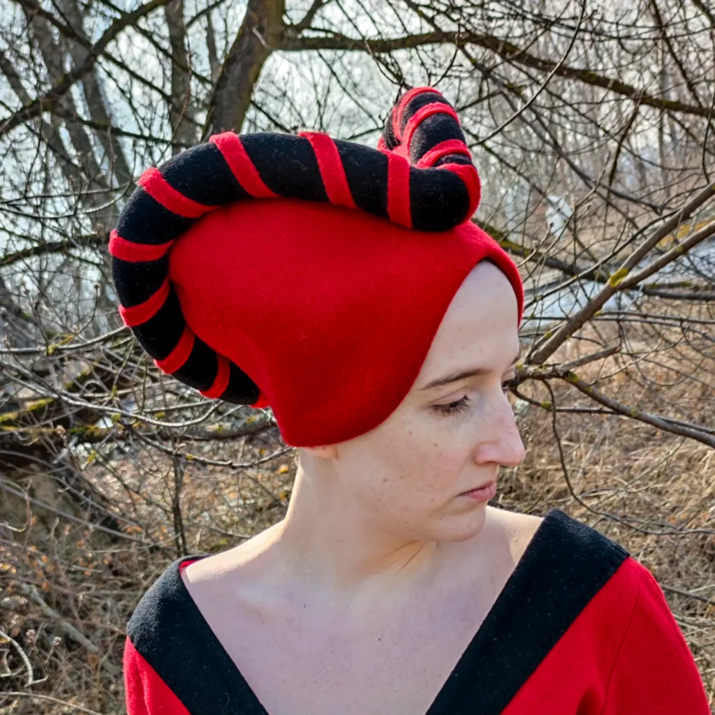 Photograph of a modern woman modeling such a hat. One can see the felt cap holds the head tightly covering the hair, but arching up high above the top of the forehead then low over the ears to make the face seem taller. A roll of black felt with a decorative wrapping of red striping it outlines a pair of horn-like curves that go symmetrically out left and right, creating almost a heart shape. Link to the maker included in the post, I love SPES Medieval Shop they've made some of my best costumes for demoing to students!