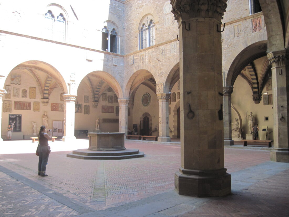 Photograph of the inside of a Medieval fortress. The interior courtyard of pale stone is surrounded by a covered loggia with rounded arches, and the walls are covered with coats of arms of past noblemen who served as captains of the police. In the center is a well, to give it is own source of water. A woman in a brown shirt stands near the well, looking very tiny in the huge courtyard.