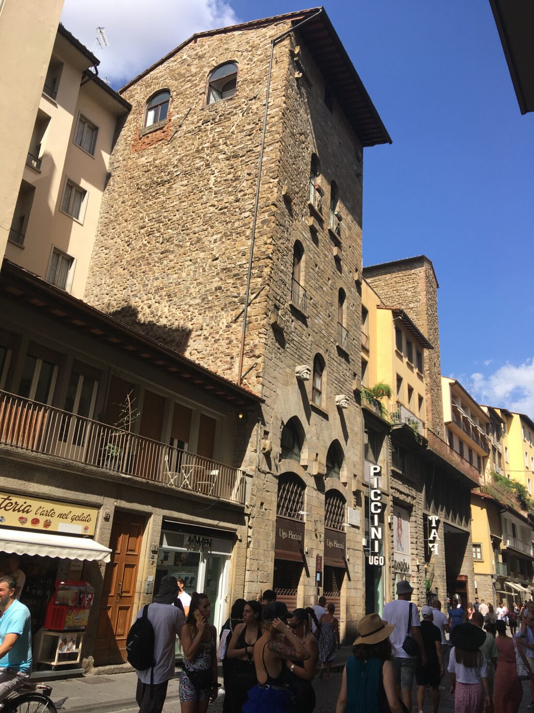 A photo of a street in Florence. Many tourists walk along and the buildings are all shops and eateries. In the center, conspicuous between buildings of yellow or beige stucco, is one building made of crude-looking yellowish stone, very rough and undecorated, with few windows and all small compared to its neighbors. A couple doors down, a second conspicuous stone section like this sticks up, also strangely blank and rough amid its yellow neighbors. Both stop about half a story above the roofs of the three-story buildings on either side of them.