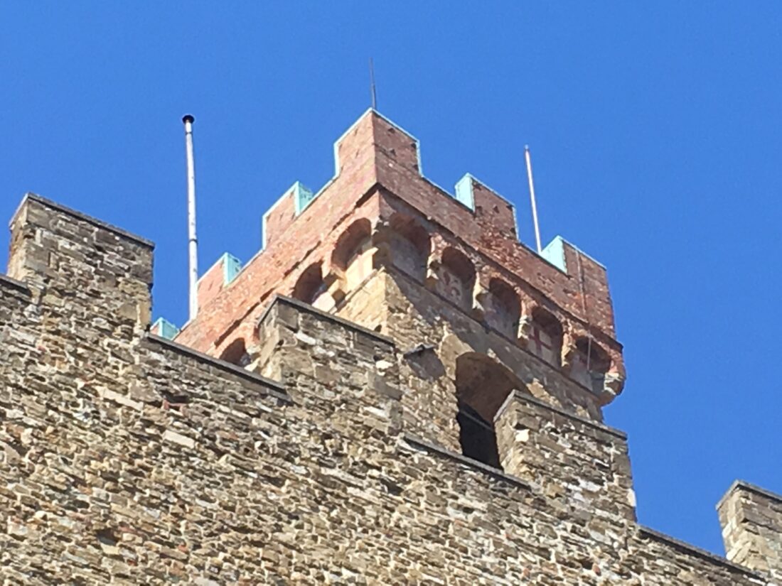 In the foreground is a stone wall with battlements. Sticking up behind it, against a bright blue sky, is the top of a stone tower. The tower itself is the same yellowish stone as the wall, but on top of the tower is a balcony area with battlements clearly added in red brick, and lined on top with metal sheeting to protect against the weather. The golden weathercock on top is on edge at the moment, and barely visible.