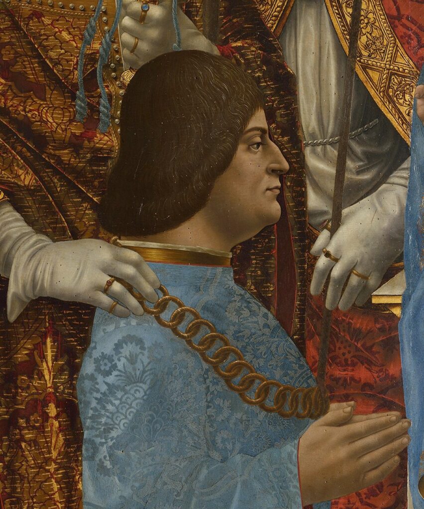 Detail portrait of Ludovico Sforza from the "Pala Sforzesca." Ludovico has black hair down to the nape of his neck, a slightly chubby build, and wears a garment of costly pomegranate pattern light blue silk brocade with gold edging and a huge gold chain around his neck. The background shows the lavish garments of the saints who surround the duke in the complete painting, which shows him and his wife amid religious figures kneeling in prayer.