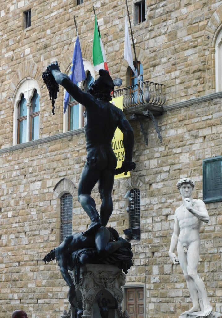 Photograph of the same bronze statue of Perseus from behind. To the lower right Michelangelo's David stands cattycorner to it, with the Medieval stone wall of Florence's Palazzo Vecchio behind it. A balcony above is crowned by the flags of the European Union, Italian Republic, and Florentine Republic.