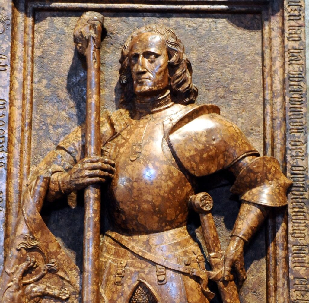 Relief sculpture in speckled orange marble from the tomb slab of a knight, Roberto Sanseverino. He is carved wearing full plate male, with a sword at his side and a spear in his hand, standing with great confidence.