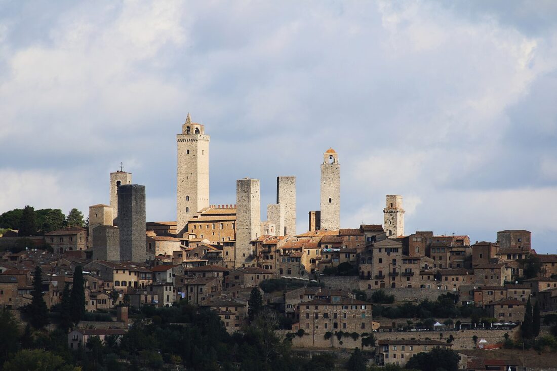 A classic image of the skyline of the town of San Gimigniano, with many smaller houses three or four stories tall with the characteristic Italian yellow stucco walls and terra cotta tiled roofs, but with eleven stone towers sticking up far above them, towering twelve stories or more. The towers are very plain and blank, just squares of stone without decoration and with few windows, clearly utilitarian more than aesthetic.