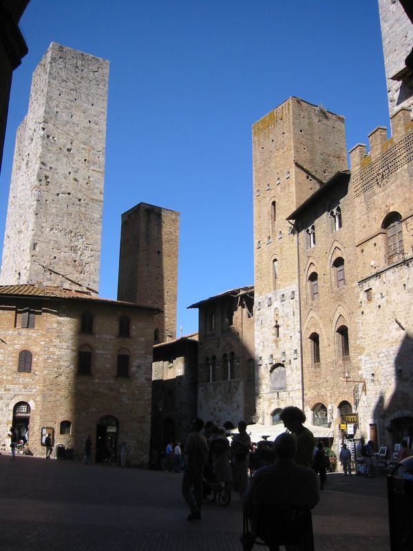 Photo of a street corner in San Gimigniano, with several plain-sided square stone towers sticking up above the roofline against a bright blue sky.