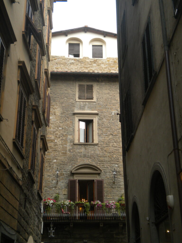 View down a street in Florence. The buildings on both sides are yellow stucco with stone windowframes, but the one at the end is naked brownish stone all the way up, with small windows one of which has a balcony outside with flowers.