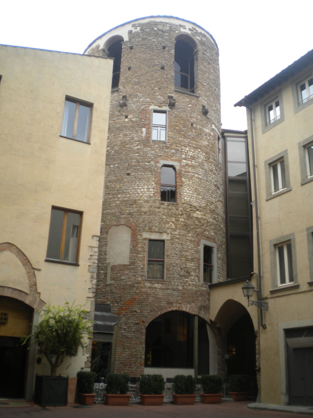 Photograph of some buildings crammed very close together. Those on the left and right are yellow stucco with large windows. In the middle, touching both of them, is a circular section made of rough nubbly stone, that really looks like it could be the bottom part of a round castle tower, it just needs battlements or a pointy cone roof. Instead it's lopped off flat just above the roofs of the other buildings.