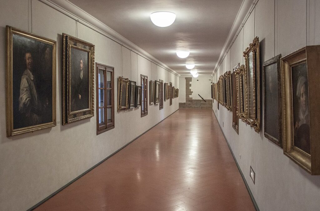 Long hallway interior, with a gently sloping floor paved with terra cotta tiles, and rows of paintings hanging along the walls.