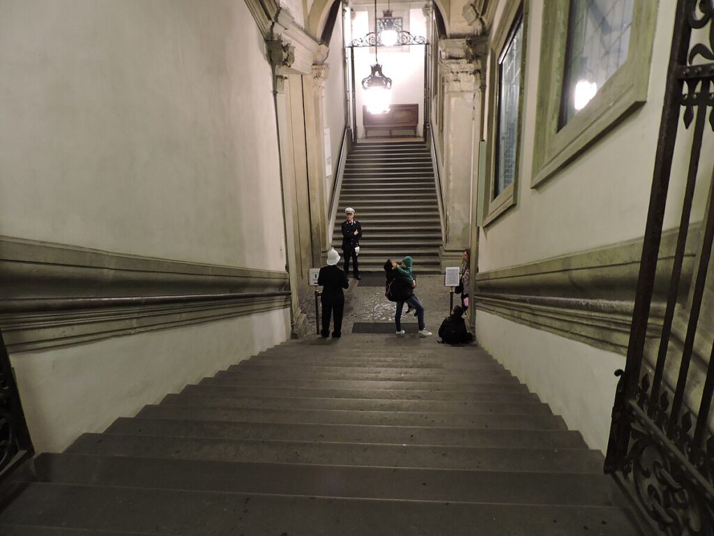 Tall, tall, tall stone staircases inside the Palazzo Vecchio, sloping intimidatingly down and up, made of slick, dark, unforgiving stone, with banisters carved into the walls. Guards stand at the bottom, and one tourist taking a photo while another tourist sits exhausted on the steps.