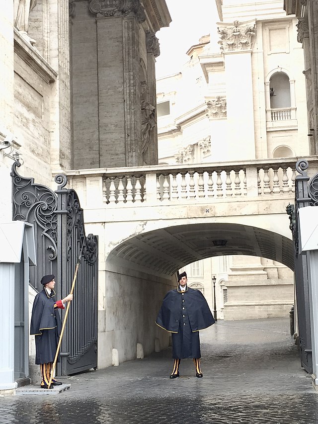 Swiss guards in uniform on duty on a cobblestone street at the Vatican. Above them is an elevated walkway above an arch, one of many elevated walkways optimizing passage without stairways throughout the maze of the Vatican, ideal for the old popes and cardinals constantly at work there. Even the modern elevators have a bench in the elevator! In the elevator! Because old guys need to sit down!