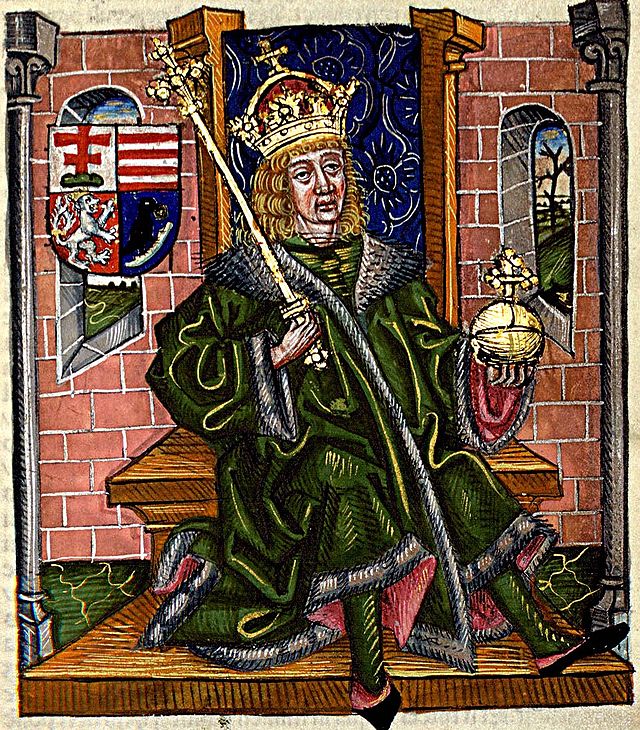 Illuminated manuscript image showing Matthias Corvinus sitting on a wooden throne, with all the trappings of power (a scepter, orb, fur-lined royal robe, and wearing the Holy Crown of Hungary, a thing he didn't actually get to do at his coronation since the ancient crown had been captured by Hungary's enemies. Behind him is a brick wall, and beside him his coat of arms. The illumination makes him look a little childlike, and he seems a bit dazed and hapless, and like he might be about to fall off his chair.